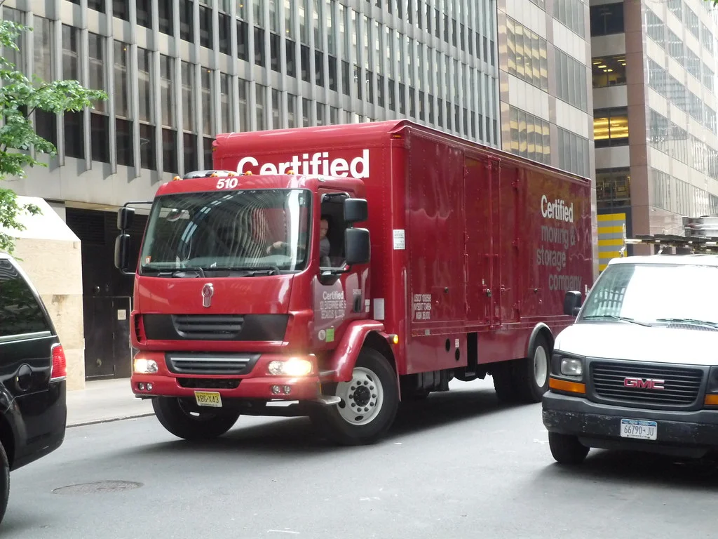 Camión de reparto rojo con carrocería de caja cerrada circulando por una calle de ciudad